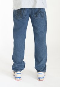 Vaqueros de mezclilla azul con un ajuste relajado y piernas rectas. Presentan costura decorativa en los bolsillos traseros. Se llevan con zapatillas blancas.