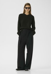 Black v-neck knit sweater paired with high-waisted black wide-leg trousers. Model wears black sunglasses and flat black shoes. Smooth texture.