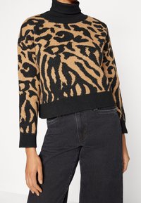 Person, der einen schwarz-beigen Pullover mit Tiermuster über einem schwarzen Rollkragenpullover trägt, kombiniert mit hochgeschnittenen schwarzen Jeans vor einem einfarbigen Hintergrund.