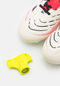 White soccer cleats with textured upper and black accents, accompanied by a bright yellow accessory tool for adjusting.