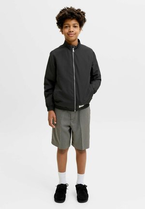 Gutt med krøllete hår iført en svart jakke med glidelås, rutete shorts, hvite sokker og svarte sko, stående mot en hvit bakgrunn.