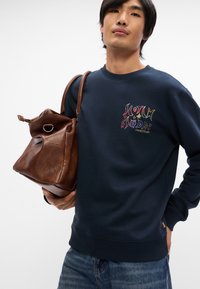 Sweatshirt bleu marine avec texte multicolore brodé sur la poitrine, associé à un sac en cuir marron avec une poignée supérieure et une boucle d'accent.