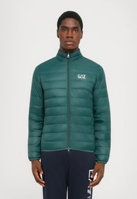Grønn pufferjakke med høy krage, horisontal quilt, frontglidelås og EA7-logo på brystet. Glatt, lett materiale.