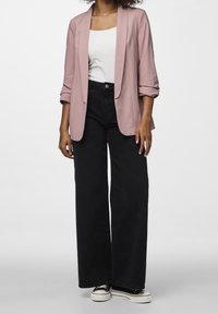 Blazer rose clair porté sur un haut blanc, associé à un pantalon large noir taille haute et des baskets noires à semelles blanches. Texture de tissu lisse.