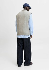 Homme portant un pull sans manches beige sur une chemise bleu rayée, un jean large noir, des chaussures marron et une casquette marine, tourné le dos.