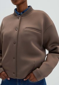 Hnědá, volná bunda s žebrovaným límcem, patentkami a náprsní kapsou. Hladká textura s moderním designem.