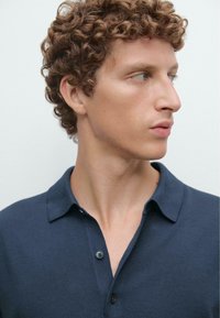 Marineblå polo-shirt lavet af blødt stof, med krave og to-knaps lukning. Modellen har kort, krøllet hår, vist i profil.