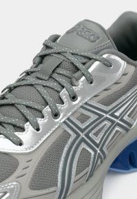 Ténis desportivo cinzento com parte superior em malha, detalhes refletivos prateados, atacadores planos e detalhe azul no calcanhar. Apresenta uma superfície texturizada e um design estruturado.