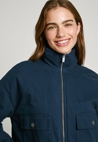 Giacca zip-up blu navy con collo alto, dotata di due tasche frontali con chiusura a pressione e un tessuto con texture.