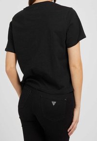 Camiseta de algodón negra con mangas cortas y cuello redondo. Presenta un logo sutil en la parte de atrás y combina con jeans de mezclilla negros con una etiqueta en forma de triángulo.