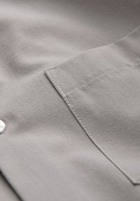 Gros plan sur un tissu gris montrant une poche de chemise cousue avec des surpiqûres soignées et un bouton blanc partiellement visible sur le côté gauche.