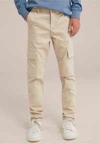 Pantaloni cargo beige con due tasche laterali, realizzati in cotone. Adattati alla caviglia, abbinati a sneaker bianche.