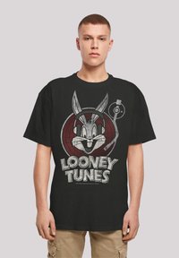 Juodas medvilninis marškinėlis su grafika, kurioje pavaizduotas Džiusas Banius su ausinėmis, įrėmintas raudoname rate. Tekstas skelbia "Looney Tunes" nusidėvėjusiu baltu šriftu.