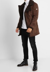 Brun, dubbelknäppt kappa med en texturerad, pälsliknande krage. Bärs över en vit polotröja, tillsammans med svarta jeans och svarta stövlar.