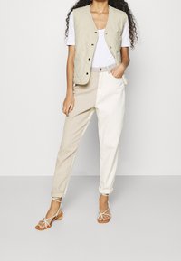 Helles beiges gesteppte Weste mit Druckknöpfen, kombiniert mit beigen und weißen schmal geschnittenen Hosen. Das Model trägt Riemchensandalen und präsentiert ein lässiges Outfit.