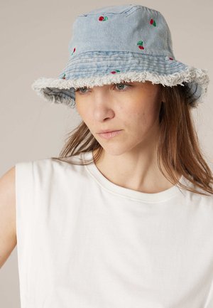 Femme portant un chapeau bob en denim bleu clair avec une broderie de cerise rouge et un bord effiloché, un haut blanc sans manches, regardant vers le bas et vers la droite.