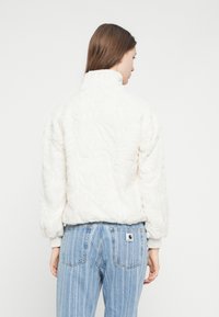 Biely, nadýchaný flísový kabátik s vysokým golierom a vzorovanou textúrou. Kombinovaný s modrými, vertikálne pruhovanými džínsami s logom na zadnom vrecku.