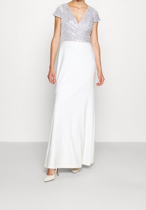 Robe longue avec un décolleté en V, corsage à sequins argentés, manches courtes, jupe blanche et tissu lisse. Associée à des chaussures beiges à talons hauts.