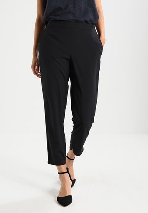 Kobieta nosząca czarne dopasowane spodnie z bocznymi kieszeniami i czarne szpilki z paskiem wokół kostki, spacerująca na tle jednolitego białego tła.