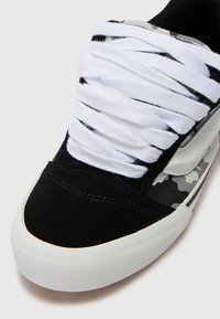 Schwarze Wildleder-Sneaker mit weißen Schnürsenkeln, ausgestattet mit einem grauen Camouflage-Seitenpanel und einer weißen Gummisohle. Glatte Textur mit kontrastierenden Materialien.