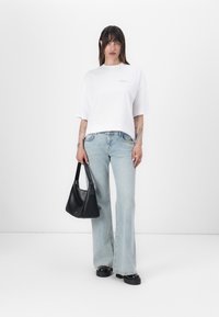 Bílá oversized T-shirt s zlatým textem, světle modré zvonové džíny, černá kabelka přes rameno a černé platformové boty, stojící na bílém pozadí.