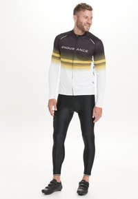 Schwarzes, langärmliges Fahrradtrikot mit weißem und verlaufendem Gelbdesign, Reißverschluss vorne und dem Text "ENDURANCE"; kombiniert mit schwarzen, figurbetonten Leggings.