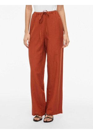 Breitbeinige, rostfarbene Hose mit Kordelzugbund, kombiniert mit schwarzen Sandalen mit goldfarbenen Akzenten an den Zehen.