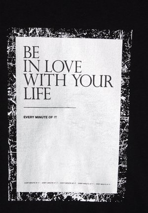 Czarna bawełniana koszulka z dużym białym prostokątnym graficznym nadrukiem z tekstem: "Kochaj swoje życie każdą minutą!" oraz postarzonym obramowaniem.