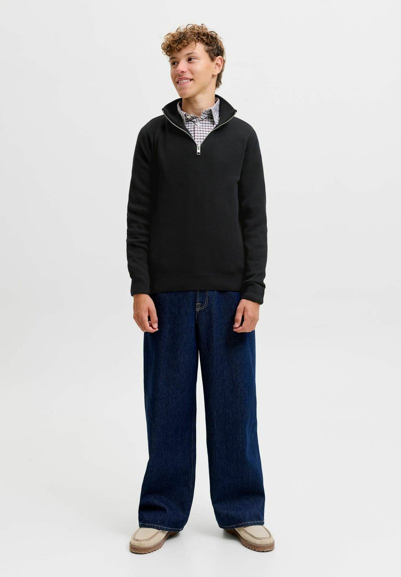 Jeune personne aux cheveux bouclés portant un pull noir à quart de zip, une chemise à carreaux, un large jean bleu et des chaussures beiges, regardant sur le côté.