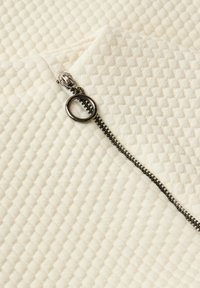 Gros plan d'une fermeture éclair en métal avec une tirette ronde sur un tissu texturé de couleur crème avec un motif en nid d'abeille.