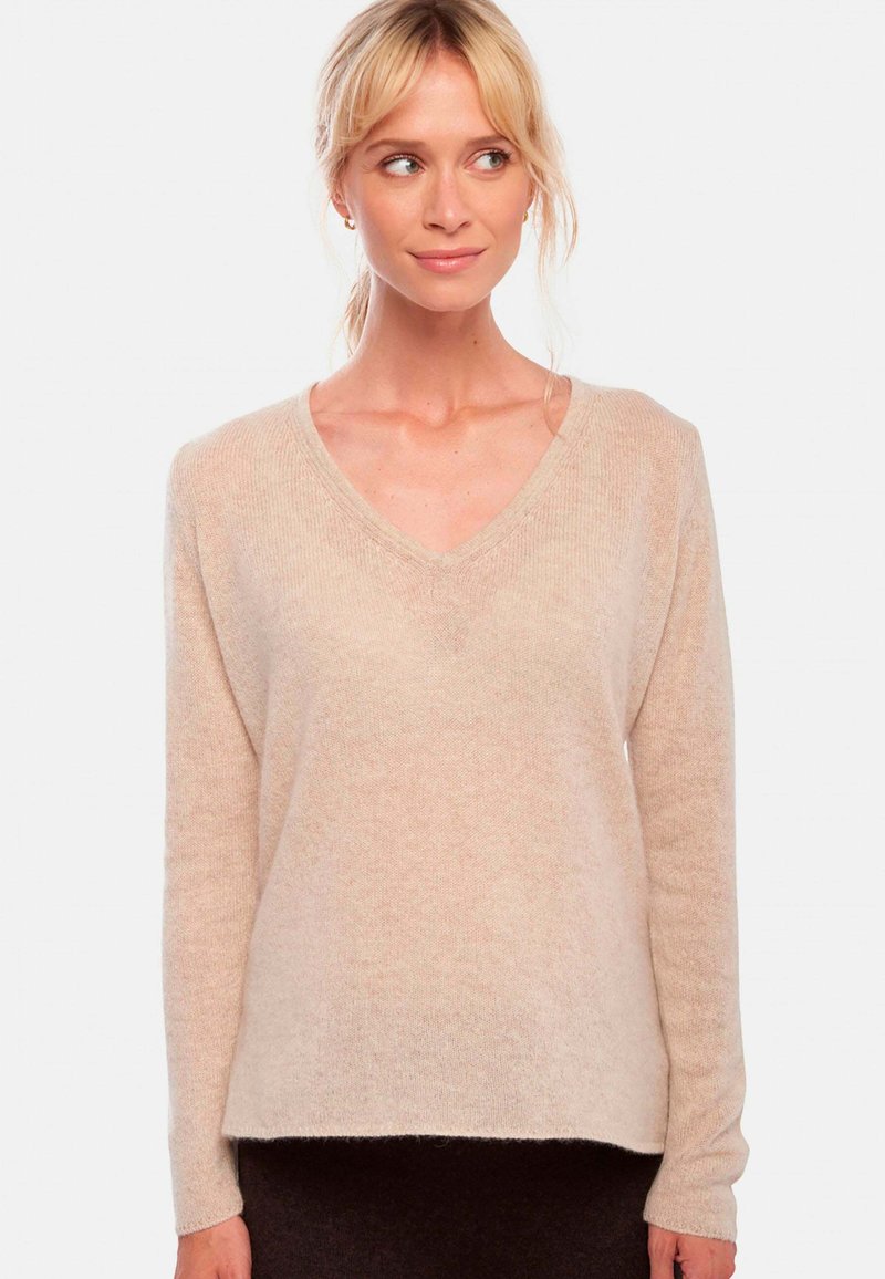 Pull à col en V, à manches longues, de couleur beige clair avec une texture tricotée douce et une coupe décontractée. Se termine légèrement au-dessus des hanches.
