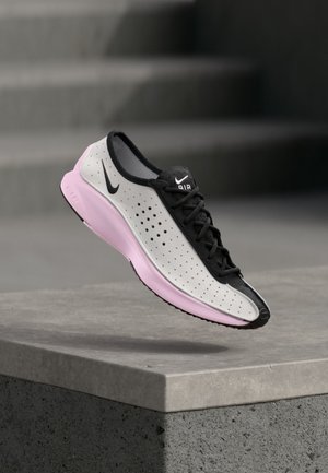 Chaussure de course Nike blanche et noire avec une semelle rose, flottant au-dessus d'un rebord en béton avec des marches d'escalier en arrière-plan flou.