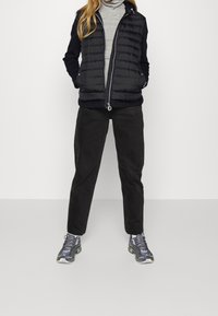 Gilet imbottito nero con cuciture verticali, dolcevita grigio, pantaloni di velluto a coste neri e sneaker grigie a fantasia.