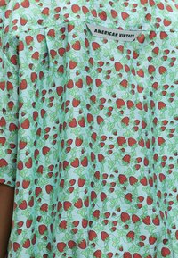 Φωτεινό γαλάζιο ύφασμα με πυκνό μοτίβο κόκκινων φράουλων και πράσινων φύλλων, με ετικέτα "American Vintage" κοντά στο λαιμό.