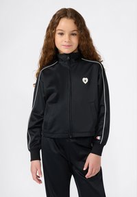 Jongeman met lang krullend haar, gekleed in een zwarte ritsjack en bijpassende broek met witte bies en logo-details.