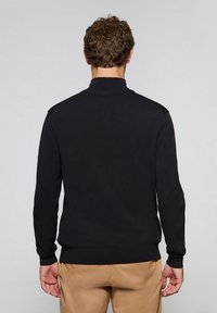 Svart tröja med lång ärm, texturerad tyg, hög krage och ribbade manschetter. Har en figursydd design och inga synliga mönster.