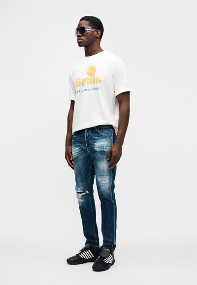 Άσπρο μπλουζάκι με κίτρινο χαμογελαστό πρόσωπο και μπλε κείμενο, ξεθωριασμένο μπλε τζιν, μαύρα παπούτσια με άσπρες ρίγες και σκούρα γυαλιά ηλίου.