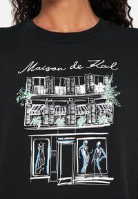 Čierna mikina s bielym a zeleným linkovým artom obchodu, s dekoratívnymi rastlinami a dvoma obrysmi šiat vo výkladných oknách.