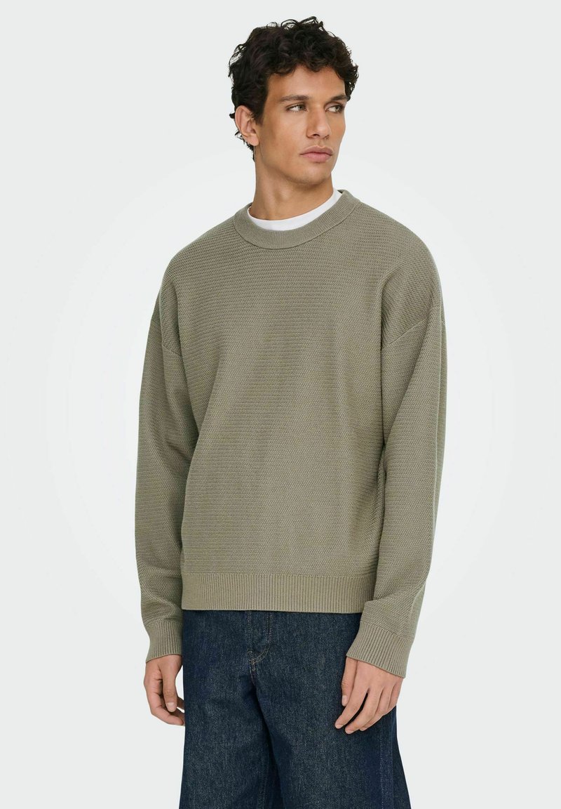 Maglione a maglia di un verde oliva chiaro con un motivo testurizzato, polsini e orlo a coste. Indossato sopra una t-shirt bianca, abbinato a pantaloni in denim blu scuro.