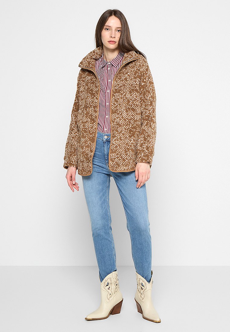 Veste en polaire brun à motifs avec un col montant, associée à une chemise rayée, un jean bleu clair et des bottes de cowboy beiges avec un détail gaufré.