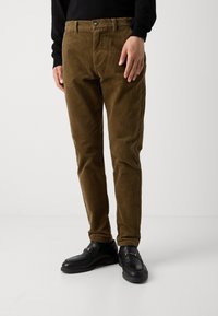 Brown corduroy pants featuring a fitted design, front pleats, and a zip fly. Paired with black loafers that have gold hardware accents.