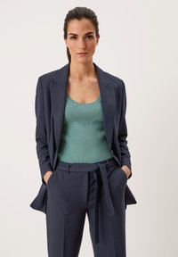 Navy blazer over een geribbelde mintgroene tanktop, op maat gemaakte broek met een gestrikt tailleband, waardoor een strakke, moderne silhouet ontstaat.