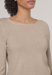 Frau trägt einen beigen Strickpullover mit einem weiten, runden Ausschnitt, lockiges Haar nach hinten gebunden und einen neutralen Lippenstift.