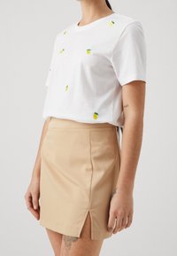 Bílý bavlněný tričko s vyšívanými žlutými ananasy, v kombinaci s béžovou mini sukni s bočním rozparkem, hladká textura.