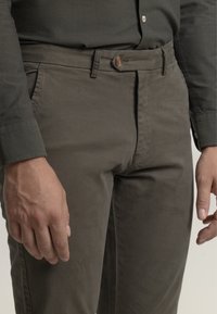 Pantalones caqui en una suave mezcla de algodón, con cierre delantero de botón, ajuste slim y bolsillos laterales. Primer plano de la cintura y la mano.