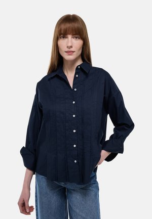 Femme aux cheveux rouges raides portant une chemise boutonnée bleu marine à manches retroussées et un jean bleu clair, devant un fond uni.