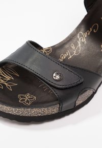 Sandale noire à bout ouvert avec bride réglable, détail bouton métal, semelle aspect liège et motif floral sur la semelle intérieure.