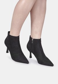 Bottes ankle en daim noir avec un bout pointu et un petit talon. Présente une fermeture à glissière sur le côté. Texture lisse avec un design épuré.