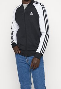 Černobílá zipová bunda s kapucí, s logem na hrudi a třemi pruhy na rukávech. Měkká tkanina, žebrované manžety.