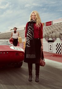 Suéter rojo, falda negra, bufanda larga negra con letras blancas, botas negras hasta la rodilla y un bolso marrón, al lado de un coche deportivo rojo.